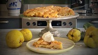Ein Stück Tarte Tatin aus Quitten ist auf einem Teller angerichtet, daneben liegen Quitten und der restliche Kuchen steht im Hintergrund