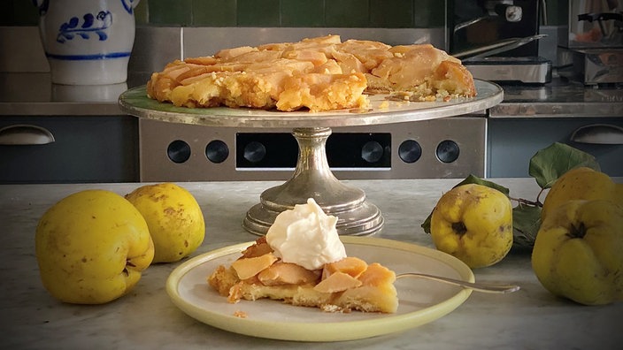 Ein Stück Tarte Tatin aus Quitten ist auf einem Teller angerichtet, daneben liegen Quitten und der restliche Kuchen steht im Hintergrund