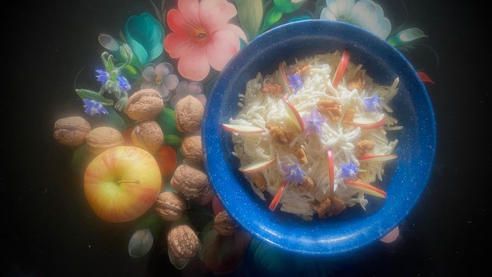 Von oben fotografiert: Eine blaue Schüssel mit Waldorfsalat auf einer Tischdecke mit Blumenmuster.Neben der Schale befinden sich ein paar Walnüsse und ein Apfel. 