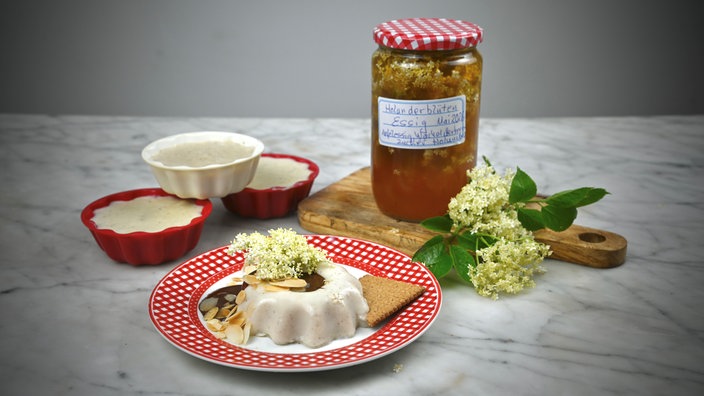 Auf einer Mamortischplatte angerichtet: Ein Teller auf dem ein Holunderblütenpudding mit einer Holunderblüte liegt, dahinter drei noch nicht gestürzte Puddingformen und ein Holzbrettchen mit einem Glas Holunderblütenessig und einer weiteren Holunderblüte.