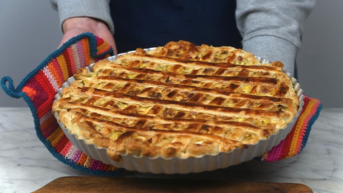 Man sieht Ullas Arme und Hände, wie sie mit bunten, gehälten Topflappen einen runden Blätterteigkuchen in die Kamera. Die Blätterteigstreifen auf dem Kuchen sind gitterförmig angeordnet.