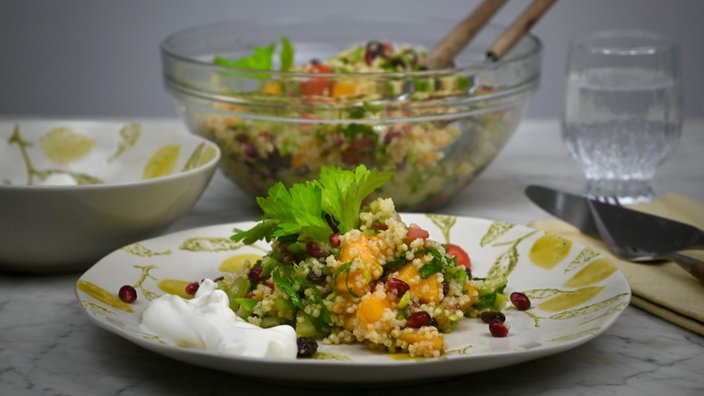 Im Vordergrund des Bildes steht ein gemustertet Teller mit etwas Hirsesalat und einem Klecks Sauce auf einem Tisch. Im Hintergrund sieht man die große Schale mit dem restlichen Hirsesalat, eine kleinere Schale mit der Sauce und ein Glas Wasser.