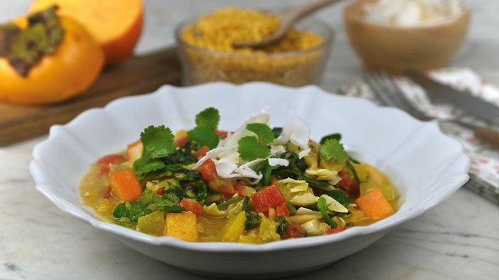 Ein weißer, tiefer Teller mit gelbem Linsencurry mit Spinat, Paprika und Kaki, garniert mit Koriander. Auf dem Tisch liegt außerdem ein Holzbrett mit aufgeschnittenen Kakis und eine Schale mit gelben Linsen.
