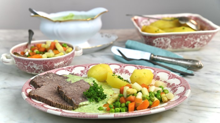 Im Vordergrund steht ein angrichteter Teller mit Rindfleisch, Kartoffeln und Leipziger Allerlei mit grüner Sauce. Im Hintergrund steht eine weitere Schale mit Leipziger Allerlei, eine Sauciere mit der Sauce, und eine Auflaufform mit Kartoffeln. 