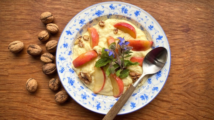 Ein tiefer, mit blauen Blumen verzierter Teller mit Grießbrei und einem Löffel steht auf einem Holztisch, daneben liegen einige Walnüsse. Der Grießbrei ist mit Apfelspalten, Walnüssen und Borretsch garniert. 