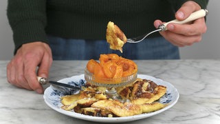 Ein Teller mit Kaiserschmarrn und einem Schälchen geröstete Aprikosen. Es wird gerade ein Stück Kaiserschmarrn mit einer Gabel vom Teller gehoben. 