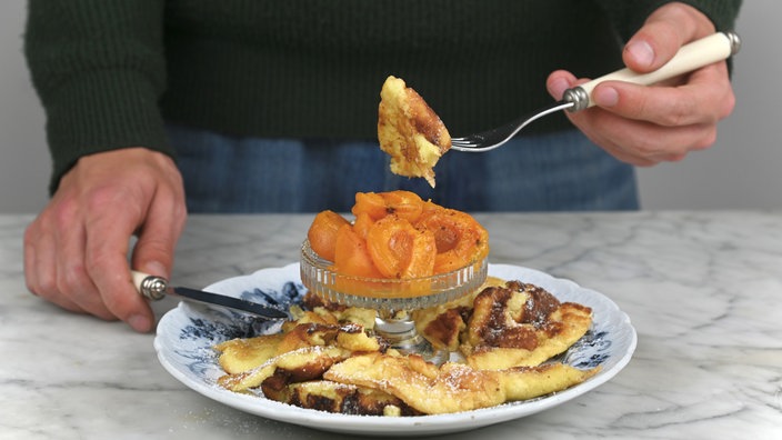 Ein Teller mit Kaiserschmarrn und einem Schälchen geröstete Aprikosen. Es wird gerade ein Stück Kaiserschmarrn mit einer Gabel vom Teller gehoben. 