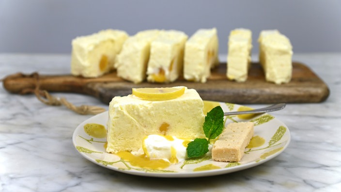 Ein Stück Lemon-Cake-Parfait auf einem Teller, garniert mit einer halben Zitrone und Minze. Im Hinterdrund stehen weitere Lemon-Cake-Parfait Stücke auf einem Holzbrett.