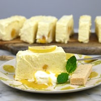 Ein Stück Lemon-Cake-Parfait auf einem Teller, garniert mit einer halben Zitrone und Minze. Im Hinterdrund stehen weitere Lemon-Cake-Parfait Stücke auf einem Holzbrett.