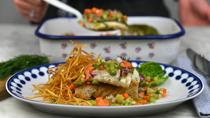 Ein angerichteter Teller mit Brot, auf dem das Stück Skrei liegt. Darüber sind Gemüsestückchen der Marinade und die Strohkartoffeln gestreut. Im Hintergrund wird aus einer Auflaufform ein weiteres Stück Skrei mit Marinade gehoben.