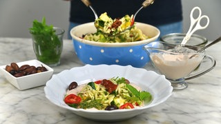Im Vordergrund steht ein tiefer Teller mit Nudelsalat, im Hintergrund sieht man die Schale mit dem restlichen Nudelsalat, aus der gerade mehr Nudelsalat gehoben wird. 
