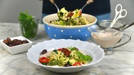 Im Vordergrund steht ein tiefer Teller mit Nudelsalat, im Hintergrund sieht man die Schale mit dem restlichen Nudelsalat, aus der gerade mehr Nudelsalat gehoben wird. 