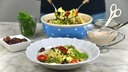 Im Vordergrund steht ein tiefer Teller mit Nudelsalat, im Hintergrund sieht man die Schale mit dem restlichen Nudelsalat, aus der gerade mehr Nudelsalat gehoben wird. 