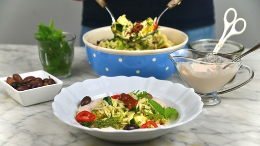 Im Vordergrund steht ein tiefer Teller mit Nudelsalat, im Hintergrund sieht man die Schale mit dem restlichen Nudelsalat, aus der gerade mehr Nudelsalat gehoben wird. 