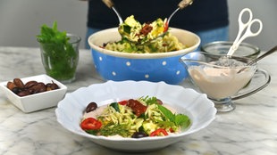 Im Vordergrund steht ein tiefer Teller mit Nudelsalat, im Hintergrund sieht man die Schale mit dem restlichen Nudelsalat, aus der gerade mehr Nudelsalat gehoben wird. 