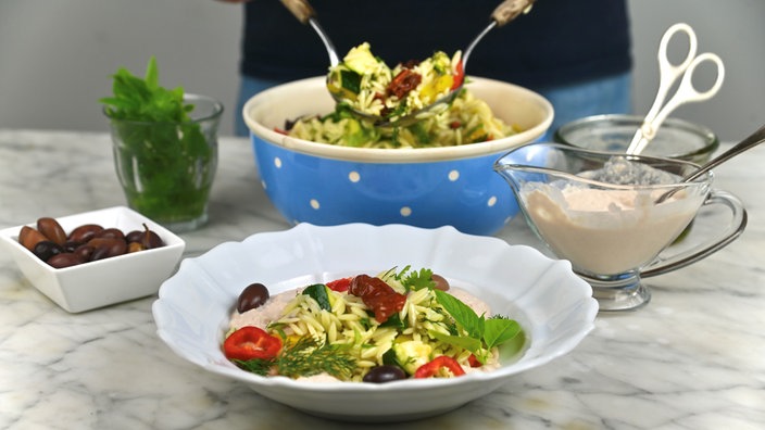 Im Vordergrund steht ein tiefer Teller mit Nudelsalat, im Hintergrund sieht man die Schale mit dem restlichen Nudelsalat, aus der gerade mehr Nudelsalat gehoben wird. 
