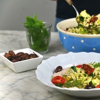 Im Vordergrund steht ein tiefer Teller mit Nudelsalat, im Hintergrund sieht man die Schale mit dem restlichen Nudelsalat, aus der gerade mehr Nudelsalat gehoben wird. 