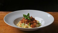 Ein weißer, tiefer Teller auf einem Holztisch mit einer schön angerichteten Portion Pasta Asciutta aus Bandnudeln, Soße und garniert mit frischem Basilikum. 
