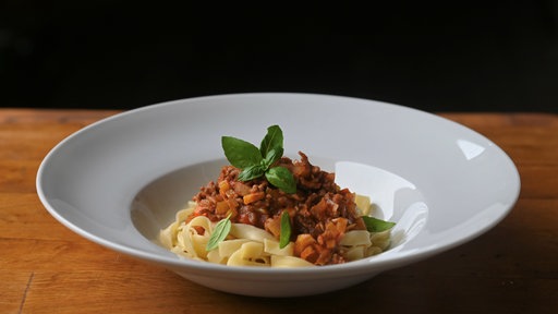 Ein weißer, tiefer Teller auf einem Holztisch mit einer schön angerichteten Portion Pasta Asciutta aus Bandnudeln, Soße und garniert mit frischem Basilikum. 