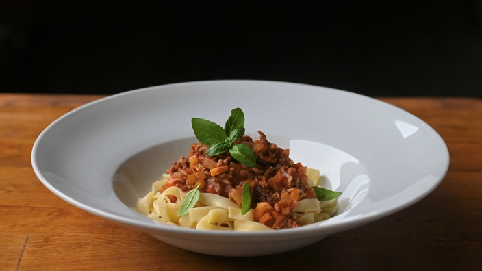 Ein weißer, tiefer Teller auf einem Holztisch mit einer schön angerichteten Portion Pasta Asciutta aus Bandnudeln, Soße und garniert mit frischem Basilikum. 