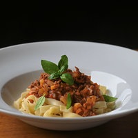 Ein weißer, tiefer Teller auf einem Holztisch mit einer schön angerichteten Portion Pasta Asciutta aus Bandnudeln, Soße und garniert mit frischem Basilikum. 