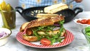 Ein rot-weißer Teller mit einem länglichen Laugen-Brioche-Brötchen mit Sesam, das mit Pesto, Schafskäse, Rucola, roten Zwieblen und Tomaten belegt ist. Im Hintergrund sieht man die restlichen Zutaten und die Pfanne mit dem Schafskäse.