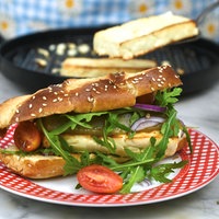 Ein rot-weißer Teller mit einem länglichen Laugen-Brioche-Brötchen mit Sesam, das mit Pesto, Schafskäse, Rucola, roten Zwieblen und Tomaten belegt ist. Im Hintergrund sieht man die restlichen Zutaten und die Pfanne mit dem Schafskäse.