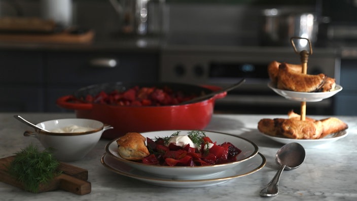 Ein elegant gedeckter Tisch: Im Vordergrund steht ein tiefer Teller mit Rote-Bete-Eintopf und einer Pirogge. Außerdem sieht man ein Holzbrettchen mit Dill, eine schale Schmand, einen roten Topf mit dem restlichen Eintopf und eine Etagere mit Piroggen.