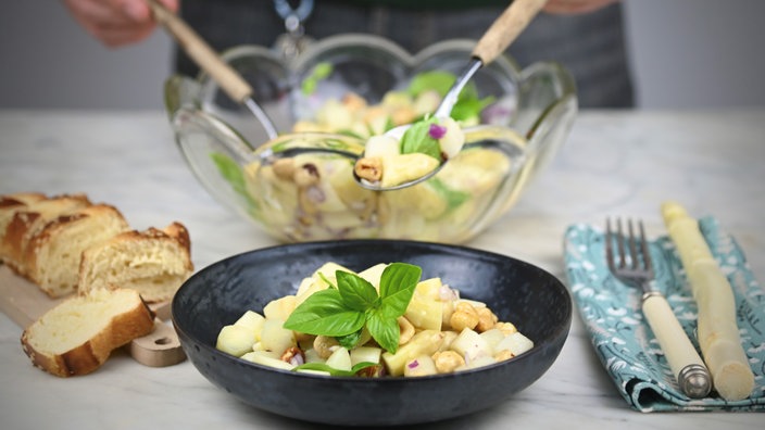 Eine angerichtete Schale Spargelsalat steht im Vordergrund. Rechts daneben liegt Besteck, links ein Brettchen mit geschnittenen Baguettestücken. Im Hintergrund sieht man die große Salatschale, aus der gerade mit Salatbesteck mehr Salat gehoben wird. 