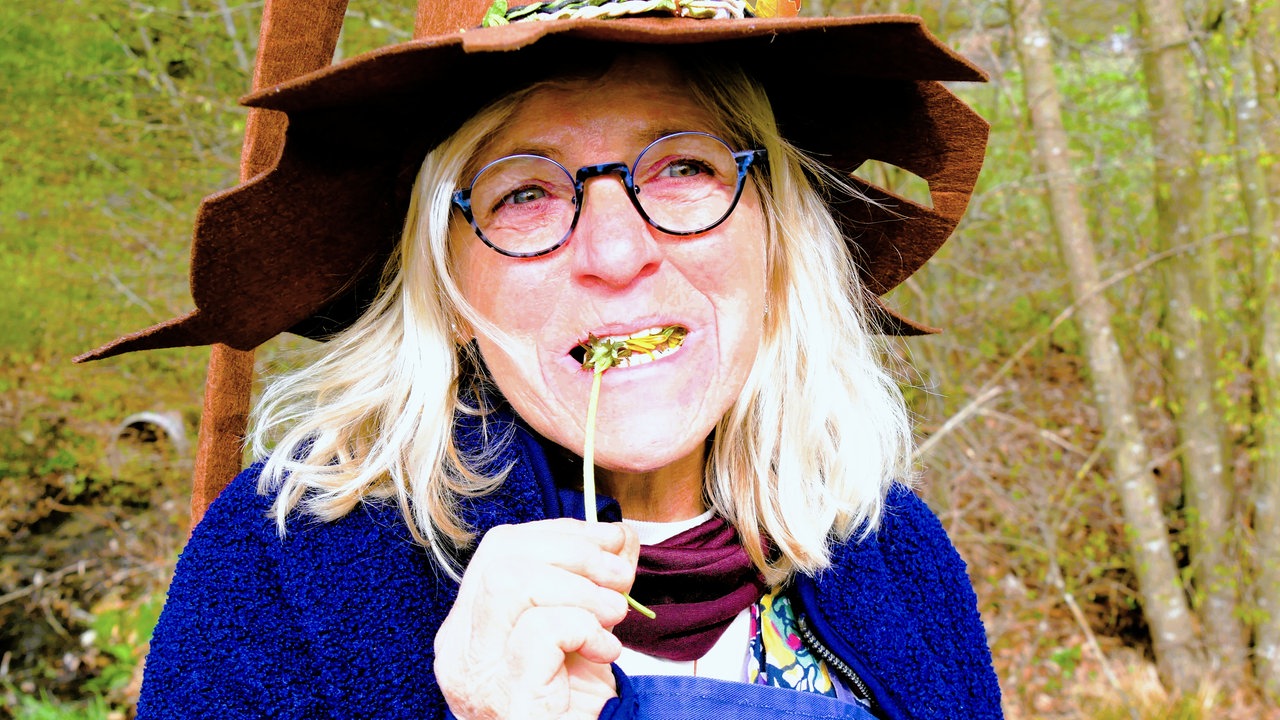 "Kraut Moni" mit Hexenhut, offenen blonden Haaren,  Brille und leuchtend blauer Jacke, beißt in einen Löwenzahn.