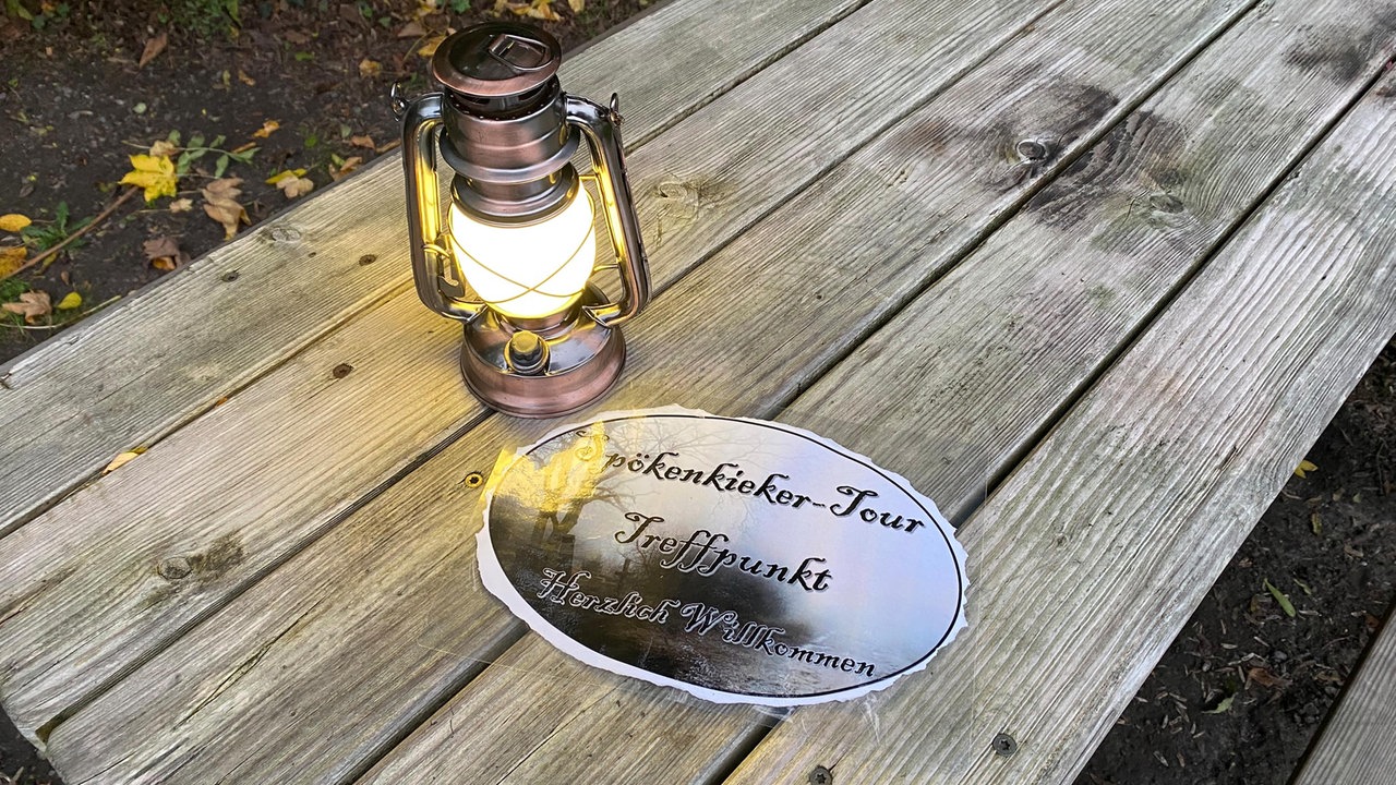 Eine Plakette mit der Aufschrift "Spökenkieker-Tour Treffpunkt. Herzlich Willkommen" und eine Laterne auf einem Holztisch.