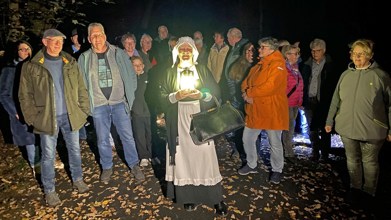 Eine verkleidete Darstellerin mit Laterne und die Teilnehmer der Wanderung im dunklen Wald.