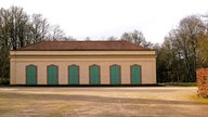Langes, helles Gebäude mit rotem Ziegeldach und mehreren grünen Türen in einer offenen Parkanlage.