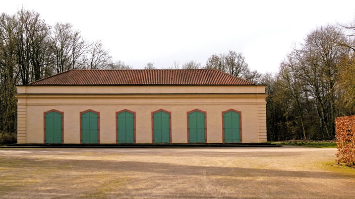 Langes, helles Gebäude mit rotem Ziegeldach und mehreren grünen Türen in einer offenen Parkanlage.