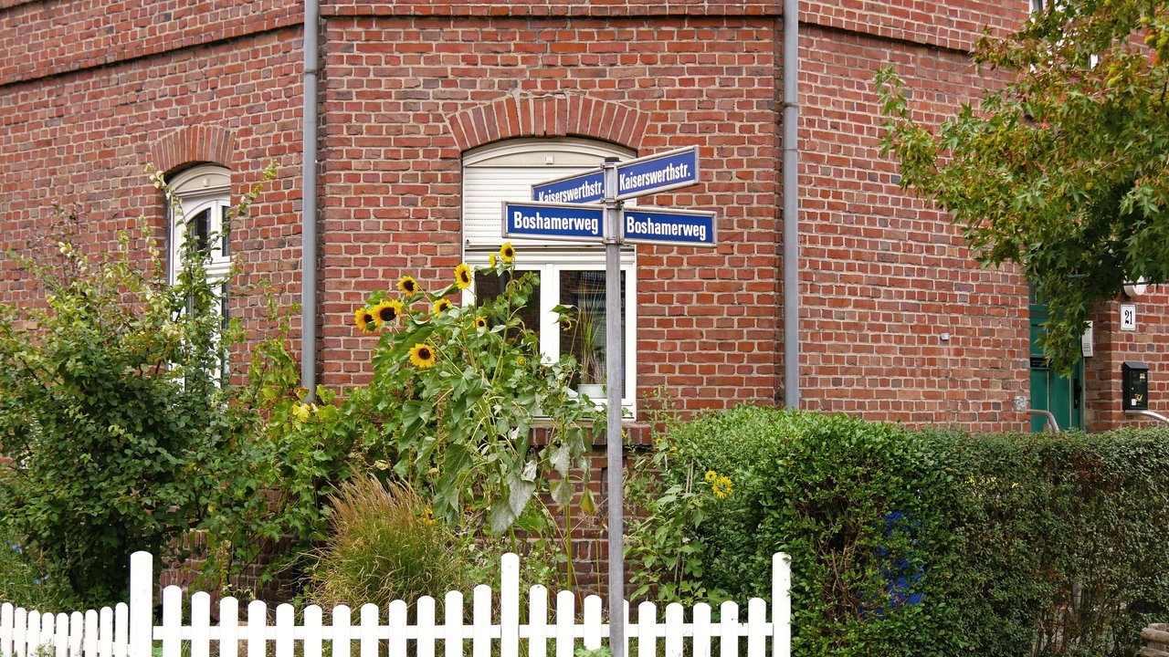 Straßenschilder, Boshamerweg und Kaiserwerthstraße, vor einem rotgeklinkertem Haus mit Sonnenblumen und weißem Zaun davor