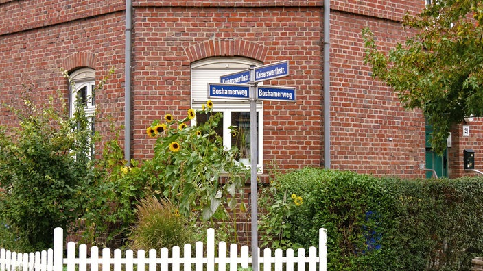 Straßenschilder, Boshamerweg und Kaiserwerthstraße, vor einem rotgeklinkertem Haus mit Sonnenblumen und weißem Zaun davor