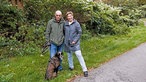 Zechenhäuser: Zwei Personen, ein Mann und eine Frau, nebeneinander, schauen in Kamera und vor ihnen ein großer Hund an der Leine, im Hintergrund Gebüsch