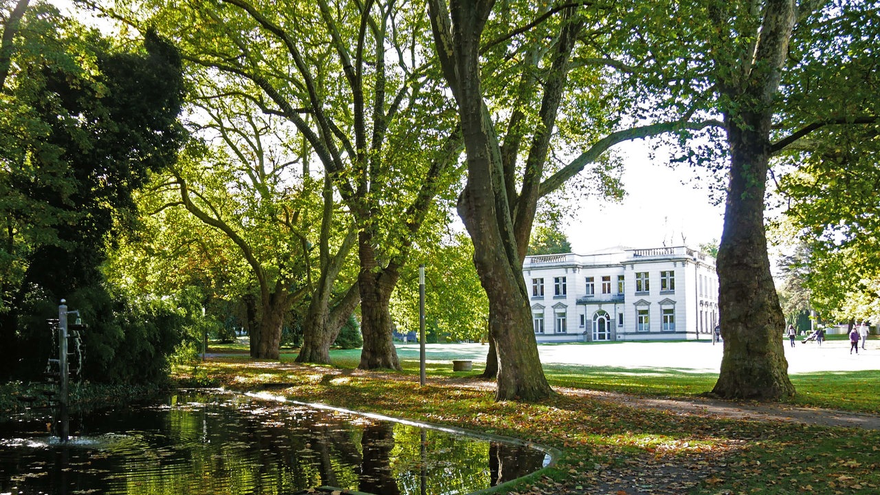 Blick in den Kurpark mit kleinem See im Vordergrund und der Kaiserpalais von Bad Oeynhausen im Hintergrund