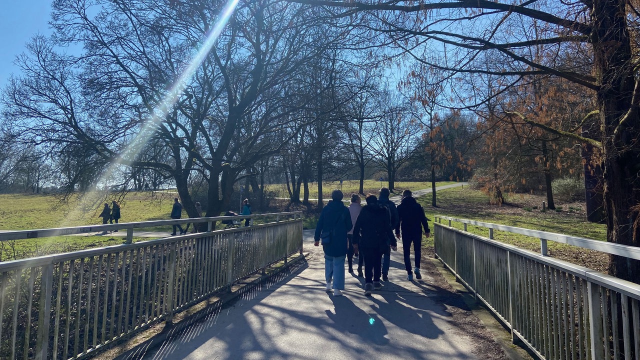 Von hinten fotografiert: Eine Gruppe Spaziergänger geht voraus, es sind Grünflächen und Bäume zu sehen. Die Sonne scheint, sodass sich ein Lichtstrahl im Bild widerspiegelt. 