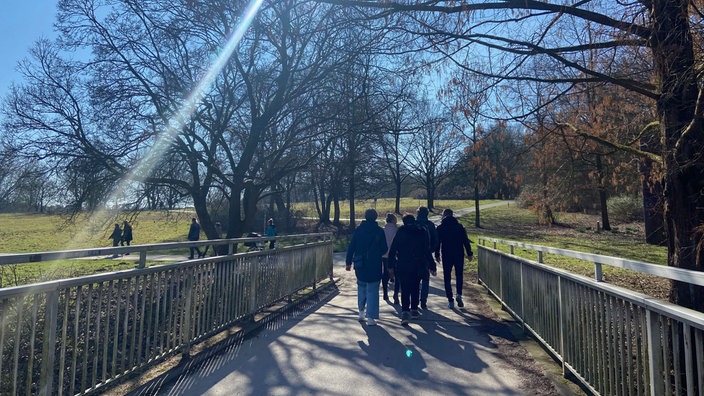 Von hinten fotografiert: Eine Gruppe Spaziergänger geht voraus, es sind Grünflächen und Bäume zu sehen. Die Sonne scheint, sodass sich ein Lichtstrahl im Bild widerspiegelt. 