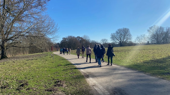 Eine Gruppe Spaziergänger von etwas weiter weg von hinten fotografiert. Rechts und lnks des Fußwegs befinden sich Rasenflächen, der Himmel ist strahlend blau. 