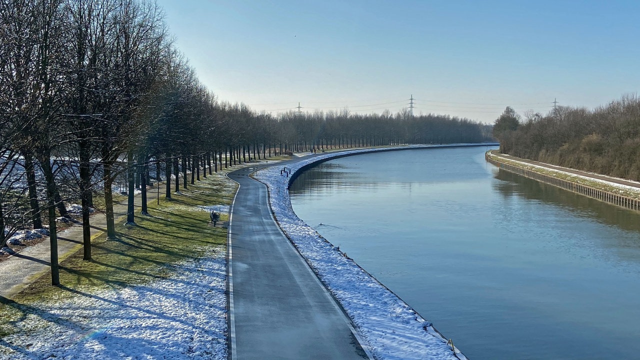 Ausblick auf den Datteln-Hamm-Kanal von oben. Am Kanal entlang gibt es einen Gehweg, der von Grünflächen und Bäumen gesäumt ist. Auf dem Bild liegt auf dem Rasen noch etwas Schnee.