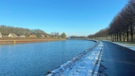 Ein Foto über den Kanal hinwegfotografiert. Auf der anderen Kanalseite ist ein Zaun zu sehen, darüber die Spitzen der Dächer kleiner Häuser.