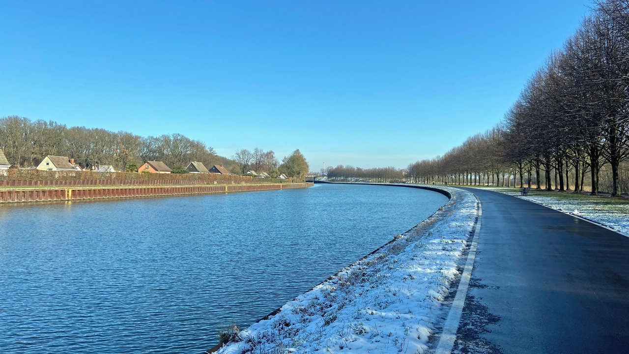 Ein Foto über den Kanal hinwegfotografiert. Auf der anderen Kanalseite ist ein Zaun zu sehen, darüber die Spitzen der Dächer kleiner Häuser.