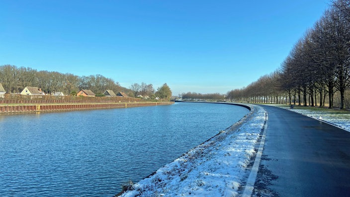 Ein Foto über den Kanal hinwegfotografiert. Auf der anderen Kanalseite ist ein Zaun zu sehen, darüber die Spitzen der Dächer kleiner Häuser.