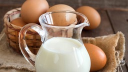 Milch in einer Kanne und Eier in einem Korb auf einem alten Holztisch. 