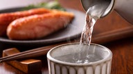 Sake wird in einen Becher geschenkt, dahinter japanisches Essen