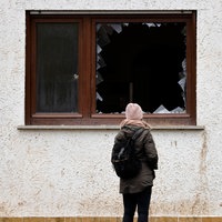 Das Beitragsbild des Dok5 Feature "Wasser bis zum Hals" zeigt eine junge Frau vor einer Hausfassade vor einem zerborstenen Fenster stehen. 