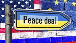 Symbolbild: Wegweiser mit Schriftzug "Peace deal" auf Nationalfarben der Ukraine vor Fahne von den USA, EU und Russland, 17.08.2025.