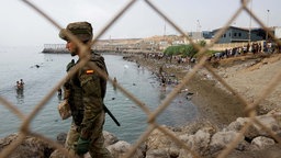 Ein Angehöriger des spanischen Militärs hält an einem Zaun Wache, während Menschen durch das Wasser waten, um nach Spanien zu gelangen